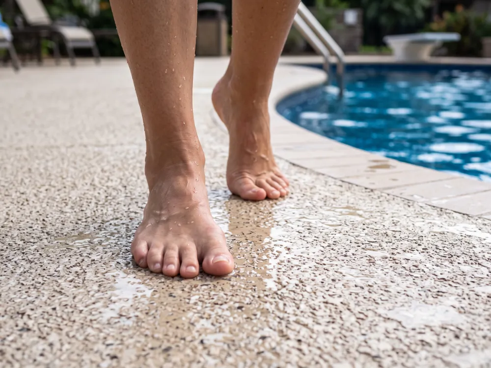 Bare feet walking on a textured light flake pool deck with water droplets near the pool edge